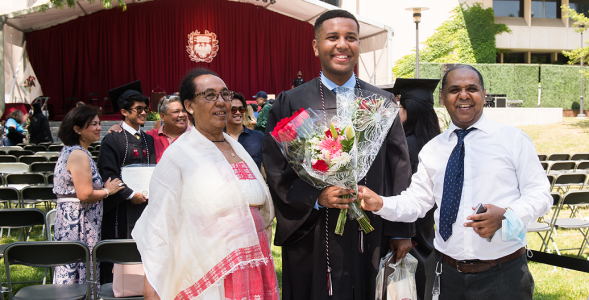 A student in a graduation gown standing with his parents holding a bouquet of flowers. 
